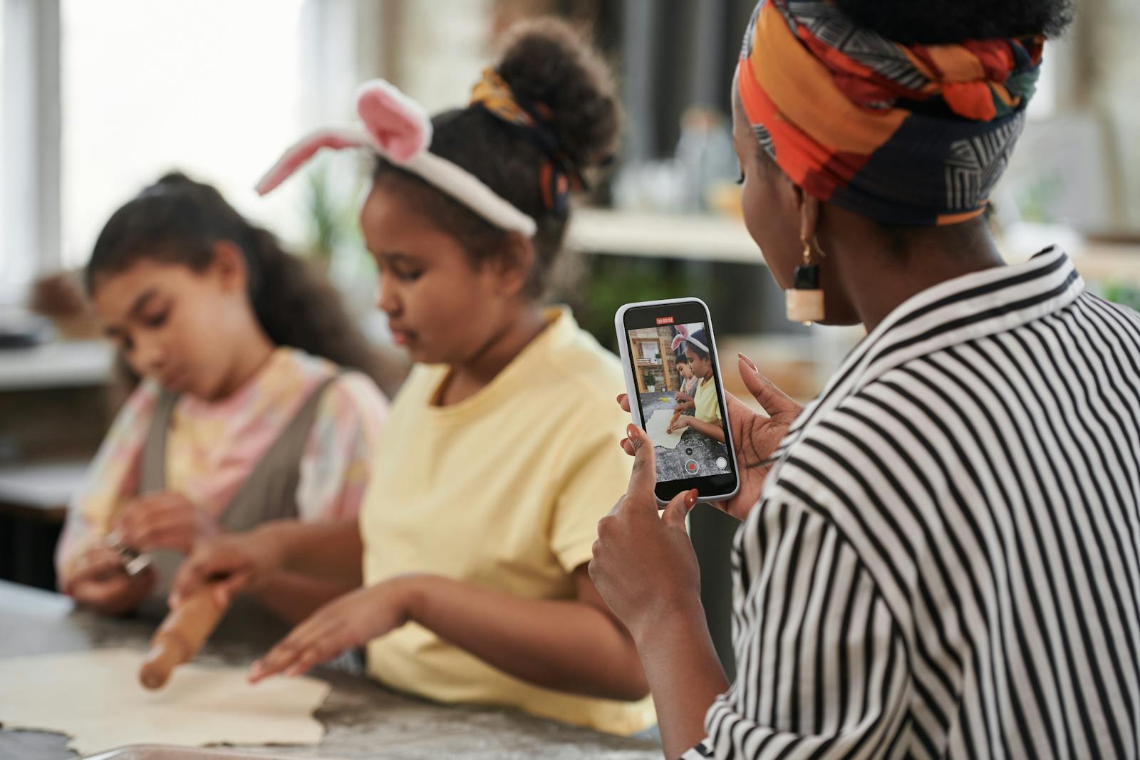 A mother records her daughters baking at home with a smartphone, capturing fun family moments.