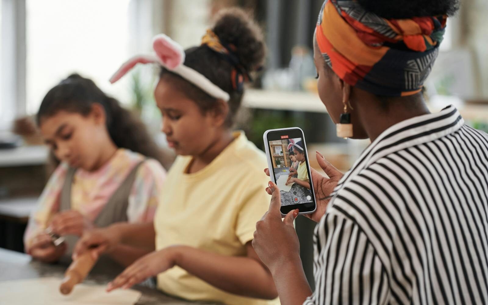 A mother records her daughters baking at home with a smartphone, capturing fun family moments.