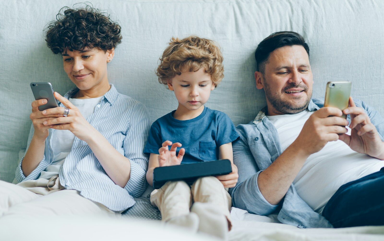 a man, woman and child sitting on a couch looking at their cell phones
